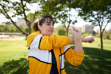 A beautiful white woman with brown hair and a sports suit with a yellow jacket prepares for exercise and exercises her muscles to avoid cramps in a park near the city wearing a Covid 19 mask     