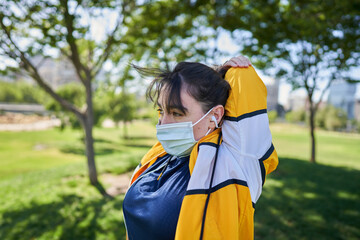 A beautiful white woman with brown hair and a sports suit with a yellow jacket prepares for exercise and exercises her muscles to avoid cramps in a park near the city wearing a Covid 19 mask     
