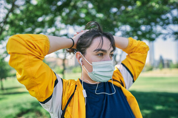 A beautiful white woman with brown hair and a sports suit with a yellow jacket prepares for exercise and exercises her muscles to avoid cramps in a park near the city wearing a Covid 19 mask     