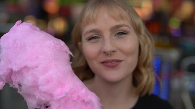 Primer plano de chica rubia con ojos azules guapa mirando a c&aacute;mara y comiendo algod&oacute;n de az&uacute;car rosa en zona de atracciones de una feria