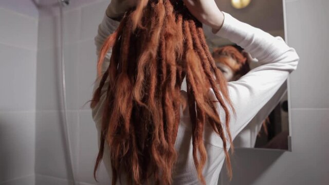 A young girl with red dreadlocks looks in the mirror and Ties dreadlocks, in bathroom in the morning getting ready during everyday routine at home