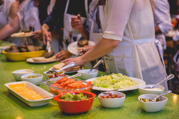 Hands of different kind of people making homemade burgers in a big green table with various ingredients (avocado, tomato, onion, mayo, etc) and drinks