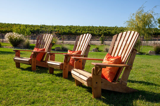 Three wooden Adirondack chairs with orange pillows sitting on the lawn with a vineyard in the background