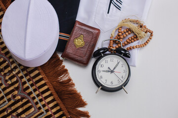 Flat lay of muslim dressed and accessories for salat with Holy Book of Al Quran and prayer beads and clock showing Duha time pray. There is Arabic word which means Holy Book