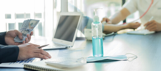 A businessman counting money and hand sanitizer on the table with a doctor examining the background