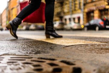 Fototapeta premium Rainy day in the big city, the woman with a red bag crosses the road on a pedestrian crossing. Close up view of a hatches at the level of the asphalt