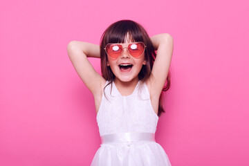 Happy small female kid wearing elegant white dress and heart-shaped glasses looks at camera with excited expressing, being glad of great event, raising hands, isolated over pink background.