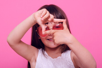 Pretty girl of preschool age imagines that she is photographer, makes a frame with her hands, looks through the cut, chooses the right angle, isolated over pink background.