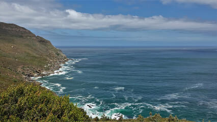 The turquoise waves of the ocean crash against the coastal stones, foaming. The rocky slopes of the hill have sparse vegetation. There are picturesque clouds in the blue sky. Cape Town. South Africa