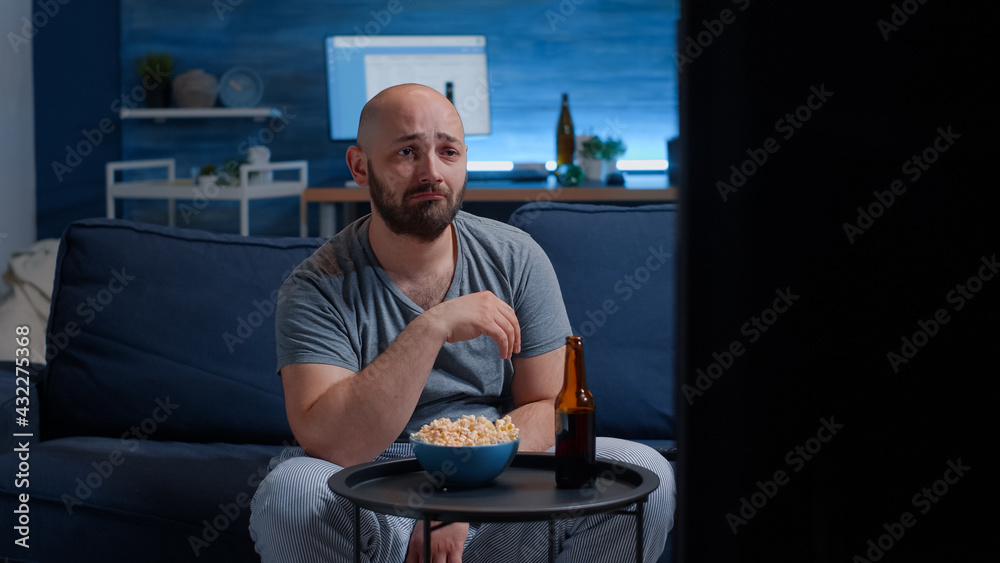 Wall mural Sad guy watching drama movie on TV crying wiping eyes sitting on couch alone at night looking at television, eating popcorn. Sensitive person affected by film action, emotionally awestruck reacting - Wall murals
