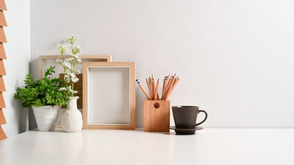 Comfortable workplace with empty frame, pencil holder, coffee cup and houseplant on white table.