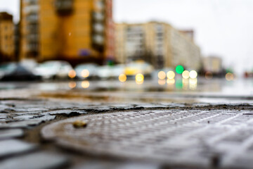 Rainy day in the big city, an empty road with glowing lights from cars. Close up view of a hatch at the level of the asphalt