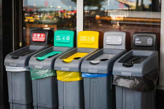 There are five trash cans on the street: red for masks, green for glass, yellow for plastic and packaging, gray for metal, and black for residual waste. Garbage sorting concept.