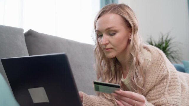 Beautiful blonde lying on the couch makes online payments with a bank card using a laptop at home. Online shopping.