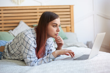 Close-up of a young woman with red hair working at a laptop lying in bed. Remote work at home.