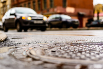 Fototapeta na wymiar Rainy day in the big city, the headlights of the approaching car on the road. Close up view of a hatch at the level of the asphalt
