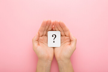 White card of question mark in young adult woman palms on pink table background. Pastel color. Point of view shot. Concept of plans, thoughts or other ideas for making decision. Closeup. Top view.