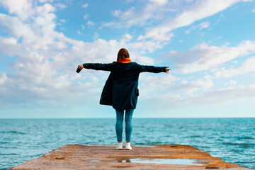 A woman in a jacket stands on the edge of the dock, her arms outstretched in flight. In the...