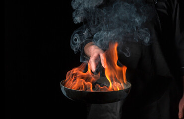  Chef cooking food in pan with fire flame on black background. Restaurant and hotel service...