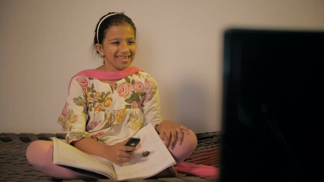 Small village girl watching her favorite program on TV while studying . Medium shot of a school-age daughter of a farmer sitting on a chaarpai and watching cartoons on TV