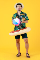 Smiling young Asian man in printed Hawaiian shirt and swim ring on waist looks at a colorful beach ball. Full body studio portrait on yellow background. Summer holiday travel preparation concept.