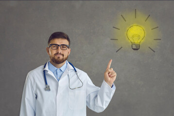 Studio portrait of proud handsome professional doctor in white lab coat pointing at yellow light bulb symbol. Concept of idea, innovation, finding new medical solutions and effective treatment methods