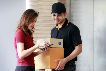 Beautiful Asian female customer signs electronic signature on mobile device while receiving packages from handsome Asian courier man. Door to door parcel shipping, e-commerce business concept.