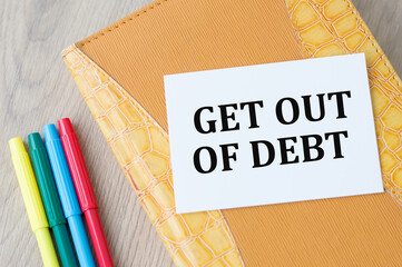 Text Get Out Of Debt on a business card that is ladyed on a notepad on a wooden background next to colored markers