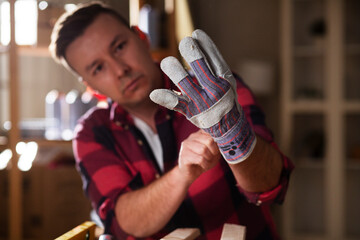 Young male carpenter working in workshop. Carpenter putting on protective gloves