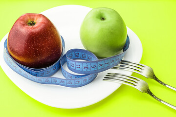 Red and yellow apples, wrapped with a centimeter tape on a white plate, on a light green background, next to two table forks. Healthy food, calorie content. Selective focus. Top and side view.