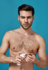 Fototapeta premium Handsome brunette naked man doing morning routine, applying foam beauty product on a cotton pad for cleansing his face, posing isolated over blue background