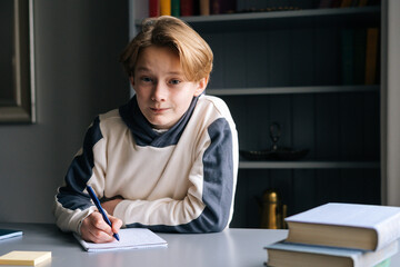 Naklejka na ściany i meble Portrait of smiling pupil boy doing homework writing to notebook with pen sitting at desk on background of book shelves and looking at camera. Child schoolboy doing homework at home.