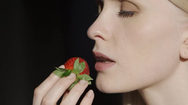 Young blond woman eating strawberries on a black background. Big ripe berry, delicious juicy strawberry. Close-up, slow motion, HD