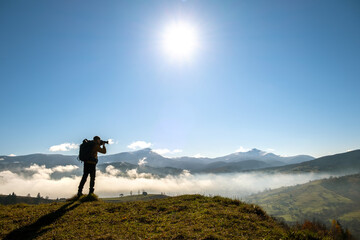 Silhouette of a backpacker photographer taking pictures of morning landscape in autumn mountains with digital camera.
