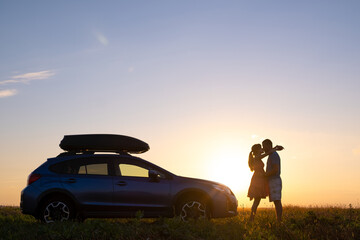 Obraz na płótnie Canvas Happy couple standing beside their SUV car during honeymoon road trip at warm summer evening. Young man and woman enjoying time together travelling by vehicle.