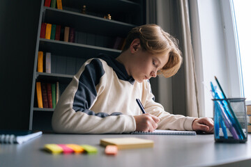 Close-up side view of thoughtful smart schoolboy making notes writing essay in notebook, sitting at desk near window at home in dark child room.