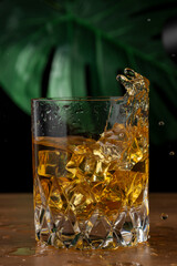 Close-up of glass with whiskey and ice in motion splashing, on wooden table, black background with illuminated green leaf, in vertical