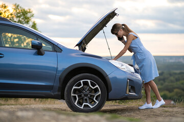 Angry female driver speaking angrily on cell phone with assistance service worker standing near a broken car with popped up hood while inspecting engine having trouble with her vehicle.