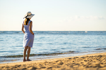 Young woman in straw hat and a dress walking alone on empty sand beach at sea shore. Lonely tourist girl looking at horizon over calm ocean surface on vacation trip.