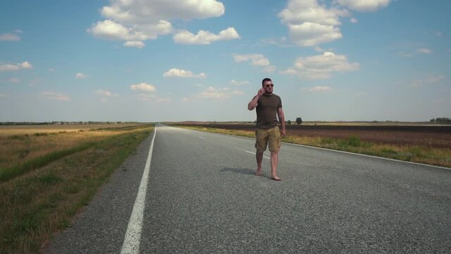 Bearded Young Man Wearing Glasses, T-shirt and Shorts Walking Barefoot on a Deserted Automobile Road Between Fields on a Hot Sunny Day. Hipster Hicker's Traveling Concept