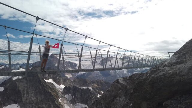 Woman with a Swiss flag on Titlis walk suspension bridge on top of Titlis mountain with glacier in the Uri Alps. Viewpoint 3028 m located in Obwalden and Bern cantons in Switzerland.