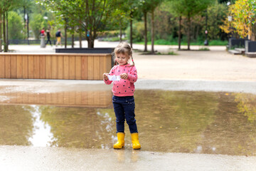 a little girl in a pink sweater and rubber boots launches a paper boat in a large puddle
