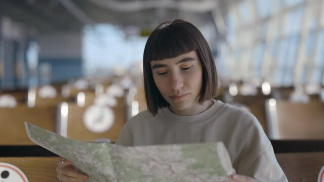 Waist up portrait of the young short haired woman sitting alone with distance at the airport and plotting the route while looking at the map