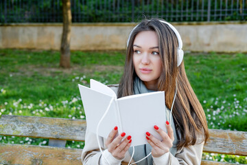 Young woman in white electronic headphones listening online audiobook in nature. Enjoying music or leisure podcast. Distance education. Storytelling