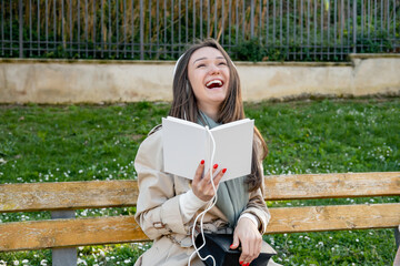 Young woman in white electronic headphones laughing and listening online audiobook in nature. Enjoying music or comic humoristic podcast. Storytelling. Leisure or spare time concept