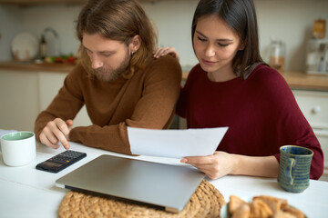 Beautiful positive young woman reading document in her hands, sitting at kitchen table with bearded husband who is calculating expenses, saving money for expensive purchase. Family and finances