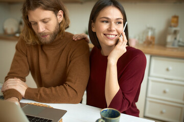 Attractive joyful young female smiling broadly, receiving good news by mobile phone, her bearded boyfriend using wireless internet connection on laptop for remote work, having focused look