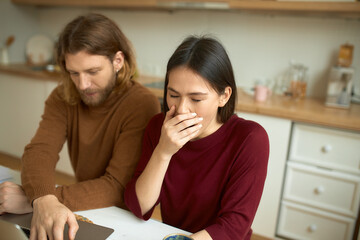 Attractive young male freelancer with beard working remotely on laptop, typing, having concentrated facial expression, his dark haired girlfriend sitting next to him, feeling tired, yawning
