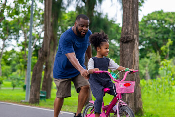 Fototapeta premium Happy mixed race family in park. African father teaching little daughter to ride a bicycle in the park. Dad and child girl kid having fun together outdoor lifestyle activity in weekend vacation.