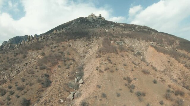 Aerial view of a summer hill slope with shrubs on blue sky with clouds on the background. Shot. Camera moving up along the steep mountain slope towards its top.